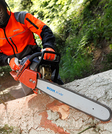 Personne en vtements de protection orange et noirs scie un tronc d’arbre abattu avec une trononneuse.