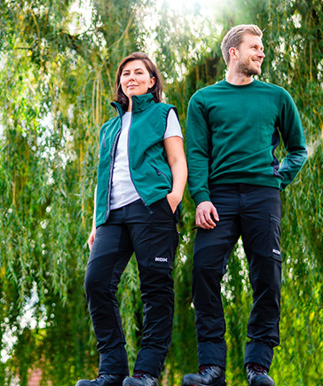 Deux personnes en vtements de jardinage verts debout cte  cte devant un fond de branches tombantes.