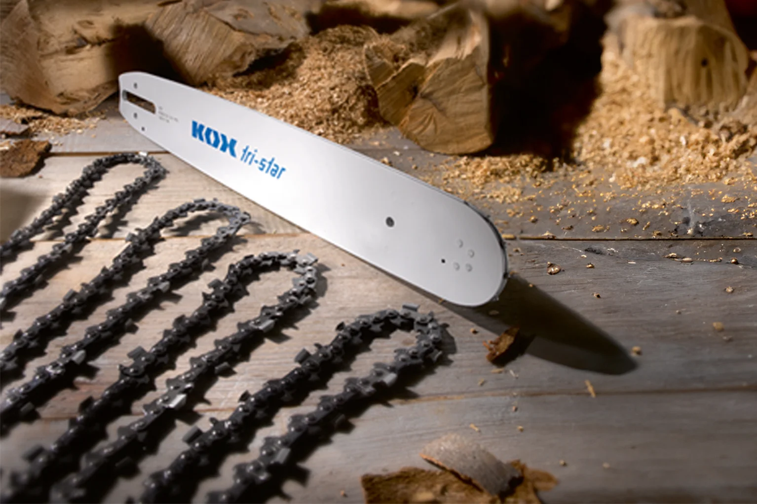 Un guide-cha�ne de tron�onneuse portant l’inscription � KOX tri-star � repose sur une surface en bois. � c�t�, plusieurs cha�nes de tron�onneuse sont dispos�es de mani�re ordonn�e, avec des morceaux de bois et de la sciure visibles en arri�re-plan.