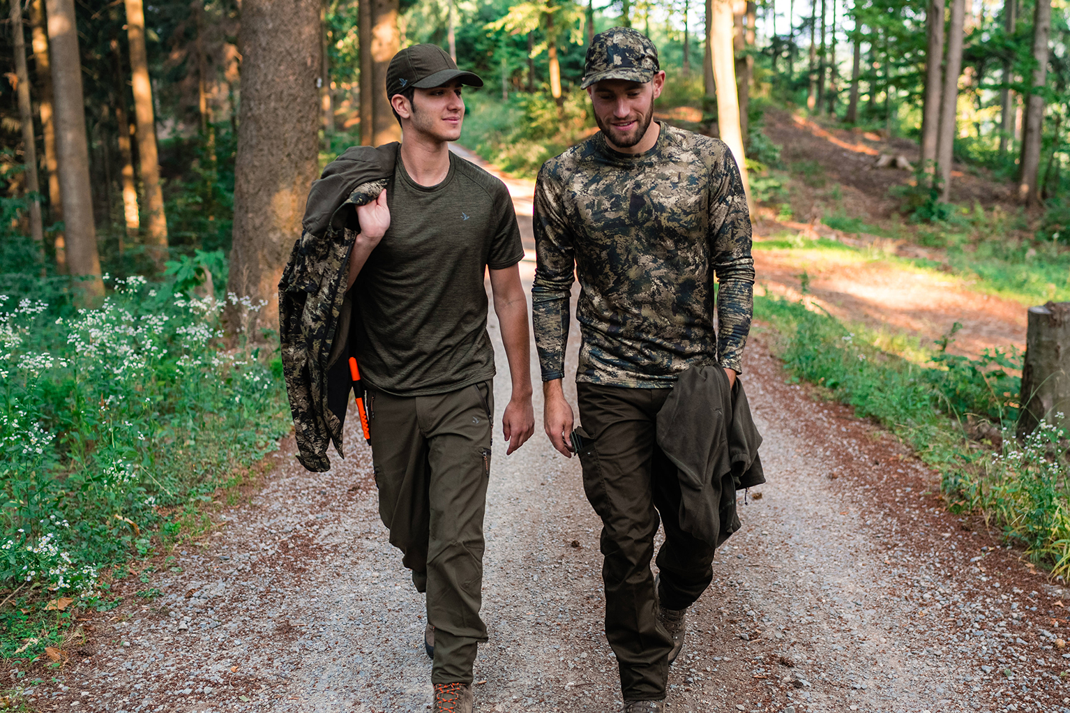 Deux hommes en tenue de chasse ou de plein air marchent c�te � c�te sur un chemin forestier ; casquettes sur la t�te, vestes � la main, en pleine conversation.