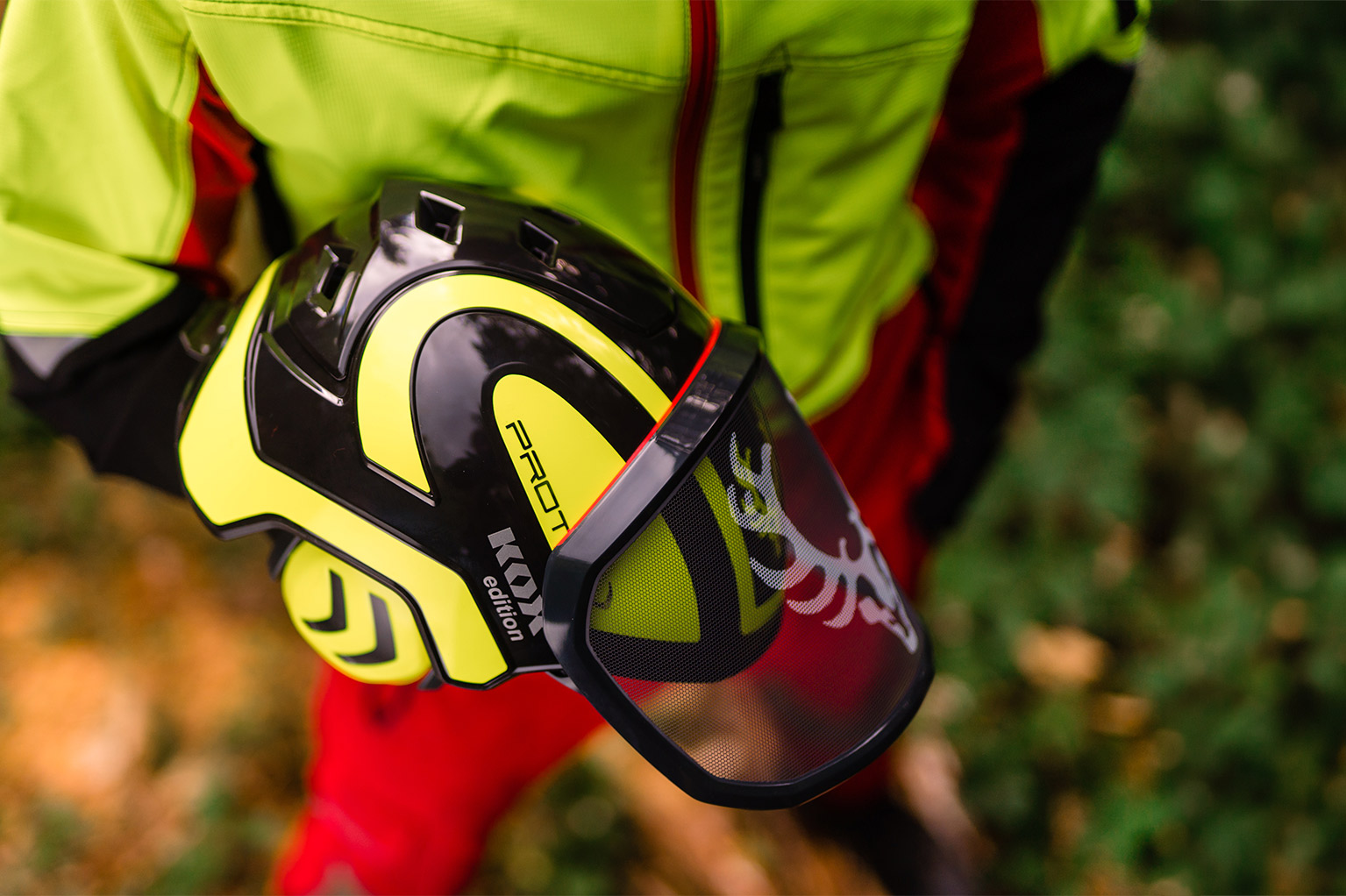 Une personne en tenue de protection jaune et rouge tient un casque forestier jaune et noir avec visi�re dans la for�t.