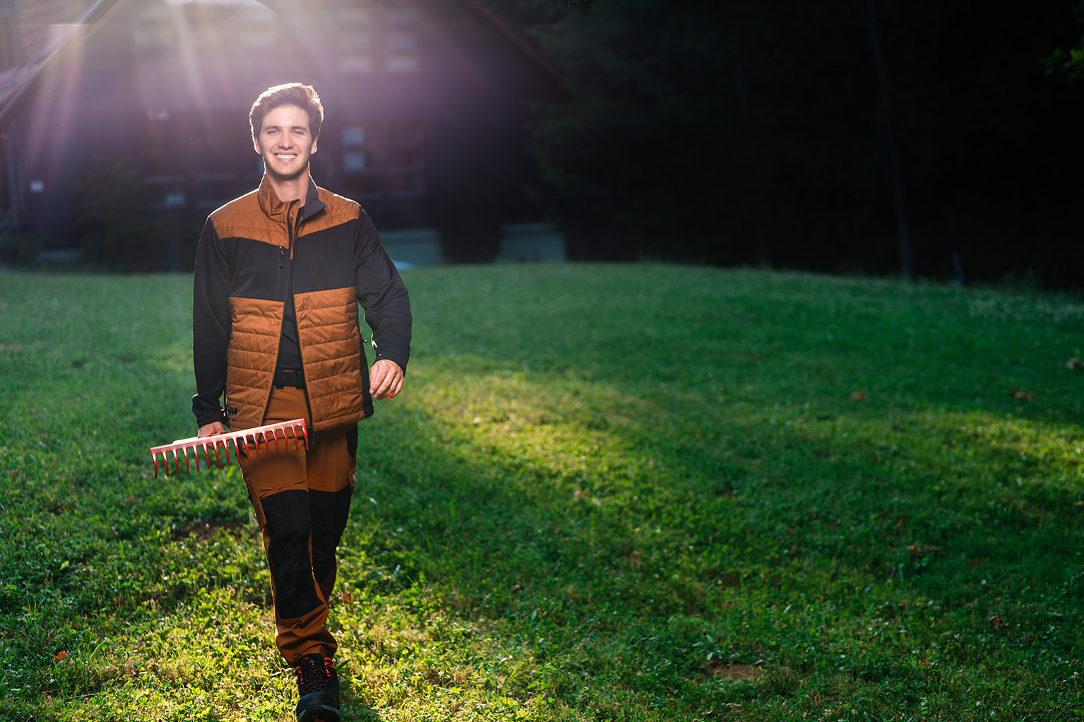 Un homme en tenue de travail marron et noire marche en souriant sur une pelouse et porte un r�teau rouge.